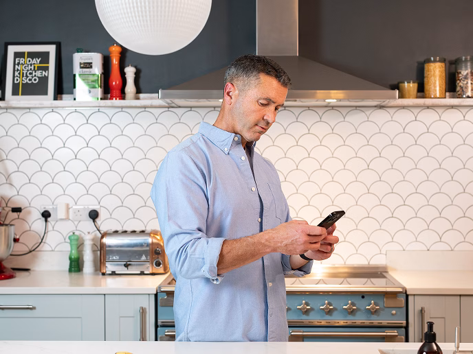 Man in a light blue shirt using a smartphone in a contemporary kitchen setting, reflecting everyday life integration with Pivotal personal subscription services.