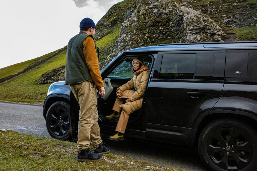 A man standing outside a black Land Rover with a woman seated inside on a countryside road, illustrating luxury car rental UK experience in a natural setting.