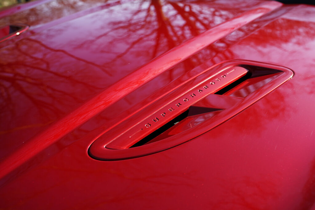 Glossy red car hood reflecting trees with a close-up of a supercharged badge air vent