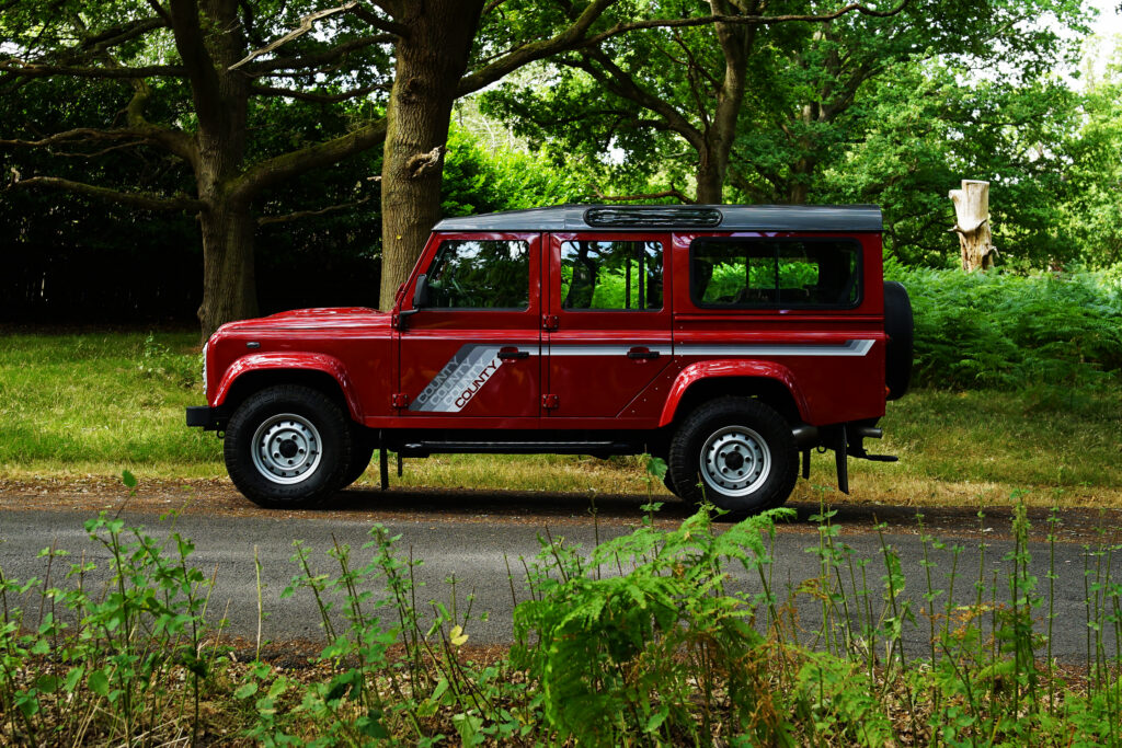 Red Land Rover Defender County with silver stripes parked on the side of a forest road surrounded by greenery and tall trees
