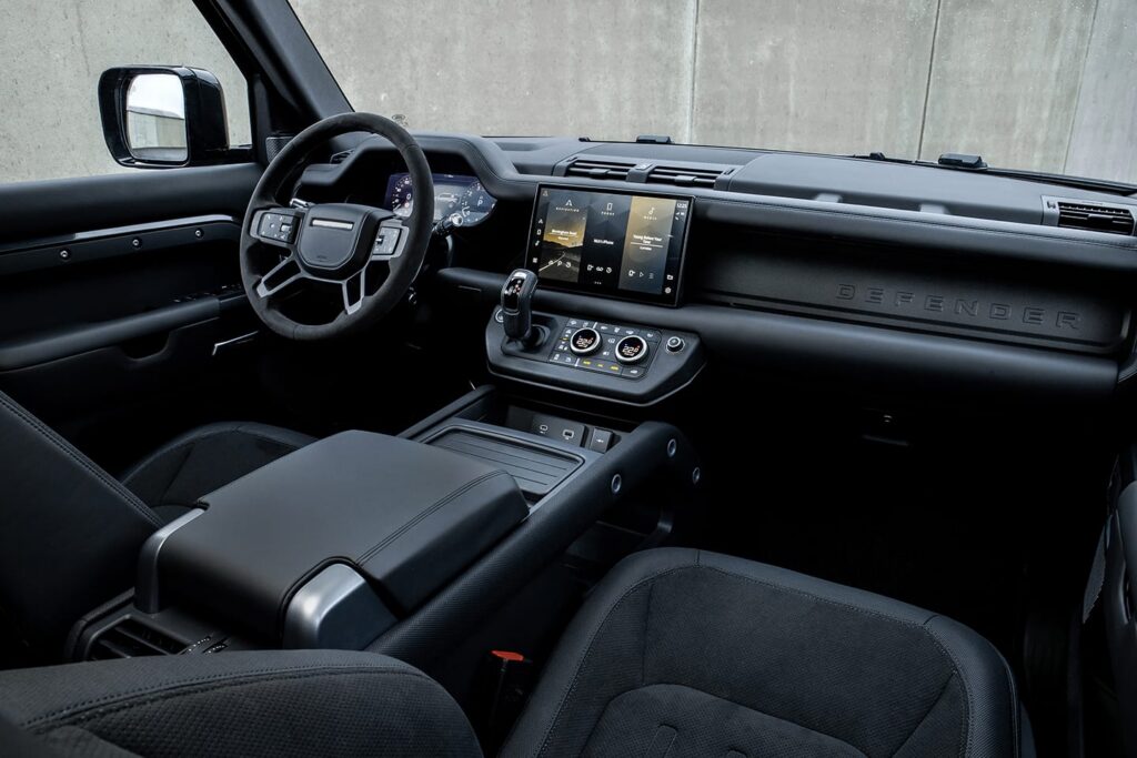 Modern black interior of a Land Rover Defender showing the steering wheel, digital instrument cluster, central touchscreen, and climate control panel.