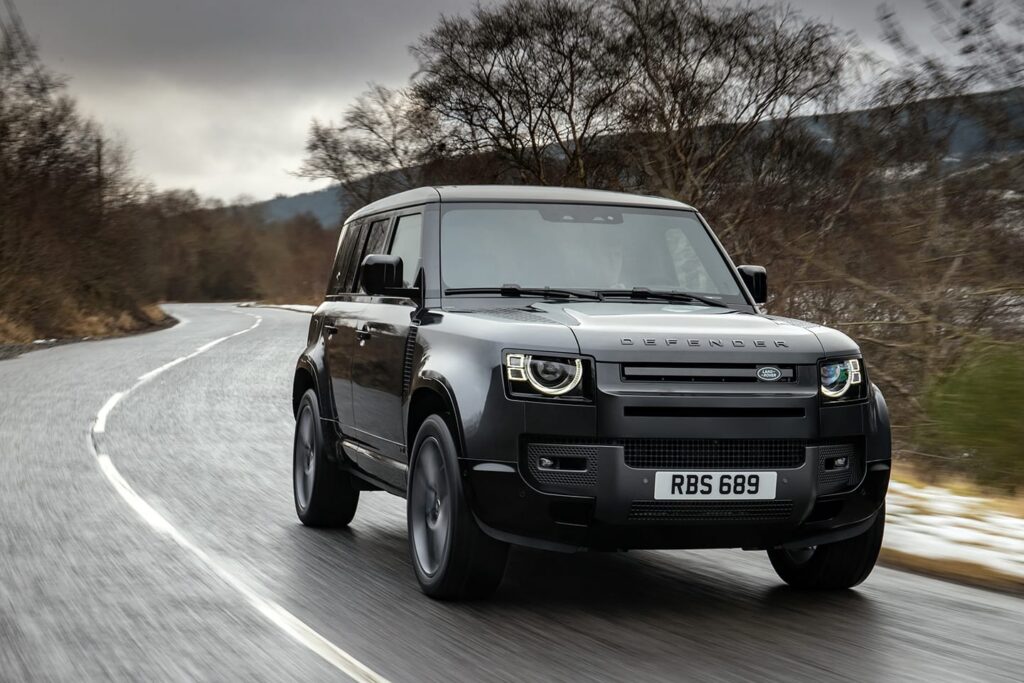 Black Land Rover Defender driving on a winding road surrounded by trees, representing luxury car rental in London.