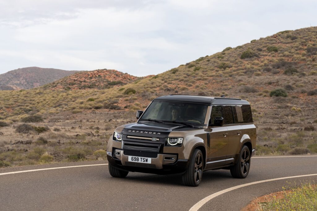 A Land Rover Defender SUV driving on a winding road through a dry, hilly landscape