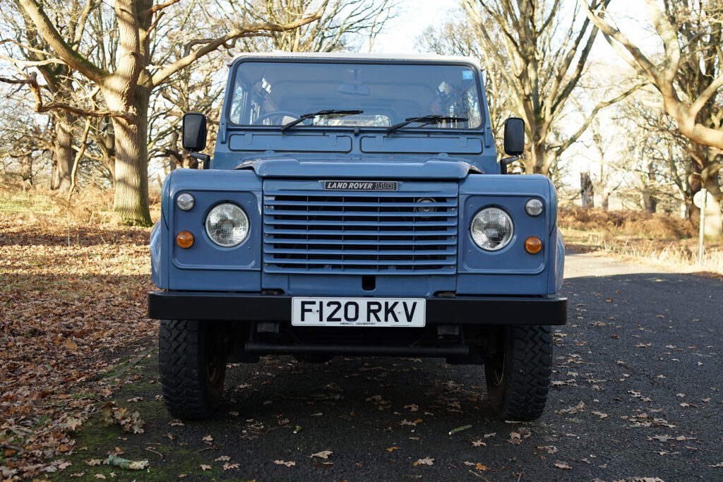 Blue Land Rover 110 parked on a leaf-strewn country road with trees in the background