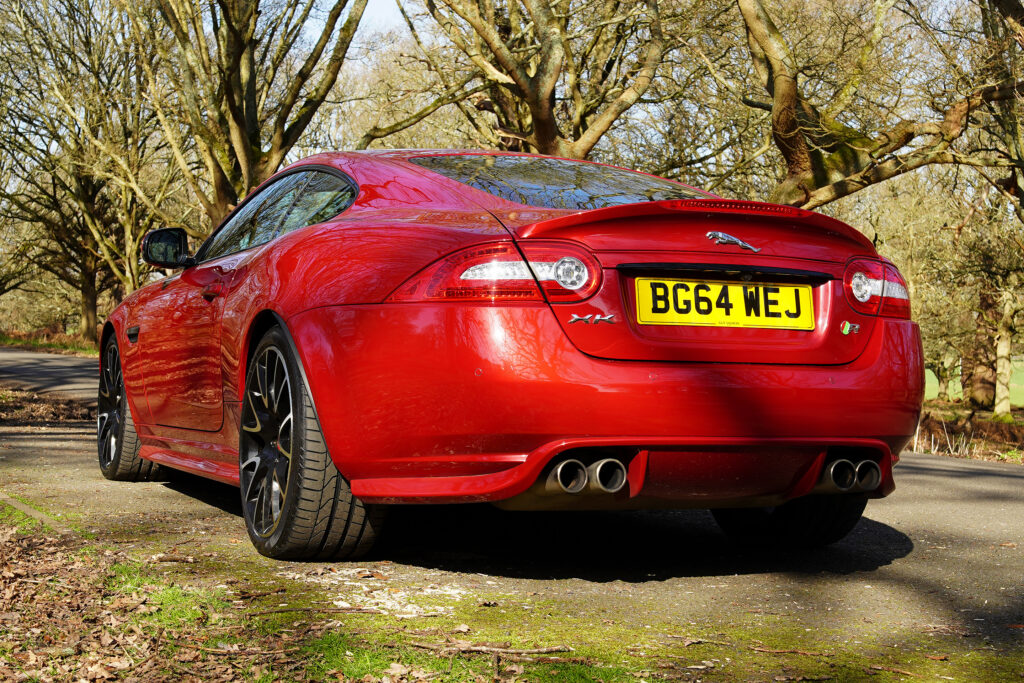 Rear view of a red Jaguar XK sports car parked on a road surrounded by trees, highlighting its tail lights and quad exhaust pipes.