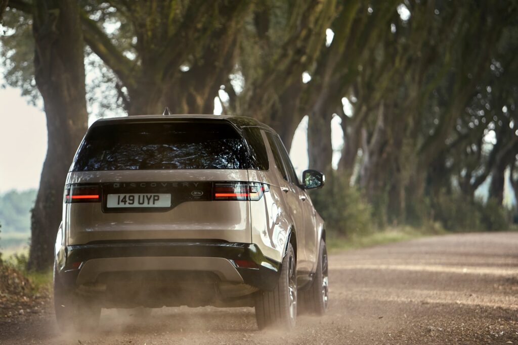 A beige Land Rover Discovery driving on a dusty tree-lined road, showcasing the vehicle's rear design and off-road capability.