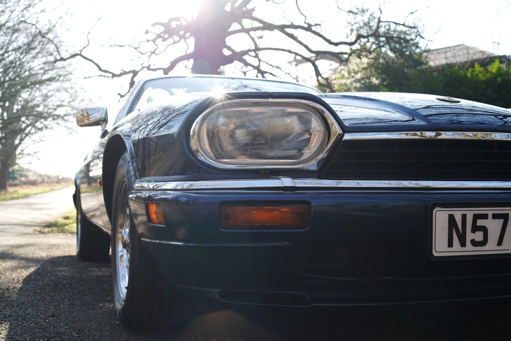 Close-up of a classic dark blue Jaguar XJS headlight and front grille on a sunny country road.