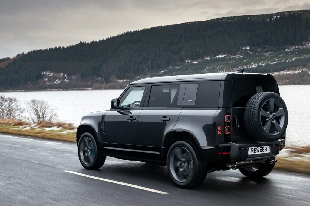 Black Land Rover Defender V8 driving along a wet road beside a lake with forested hills in the background