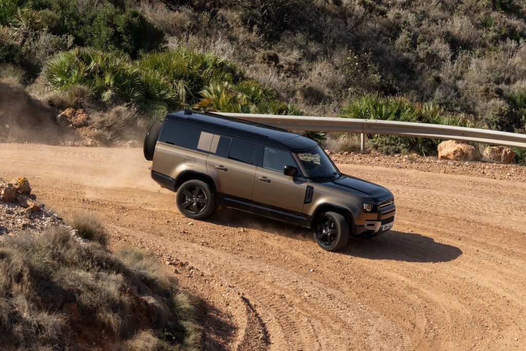 A Land Rover Defender 130 driving on a winding dirt road with greenery on the sides in a rugged terrain setting.