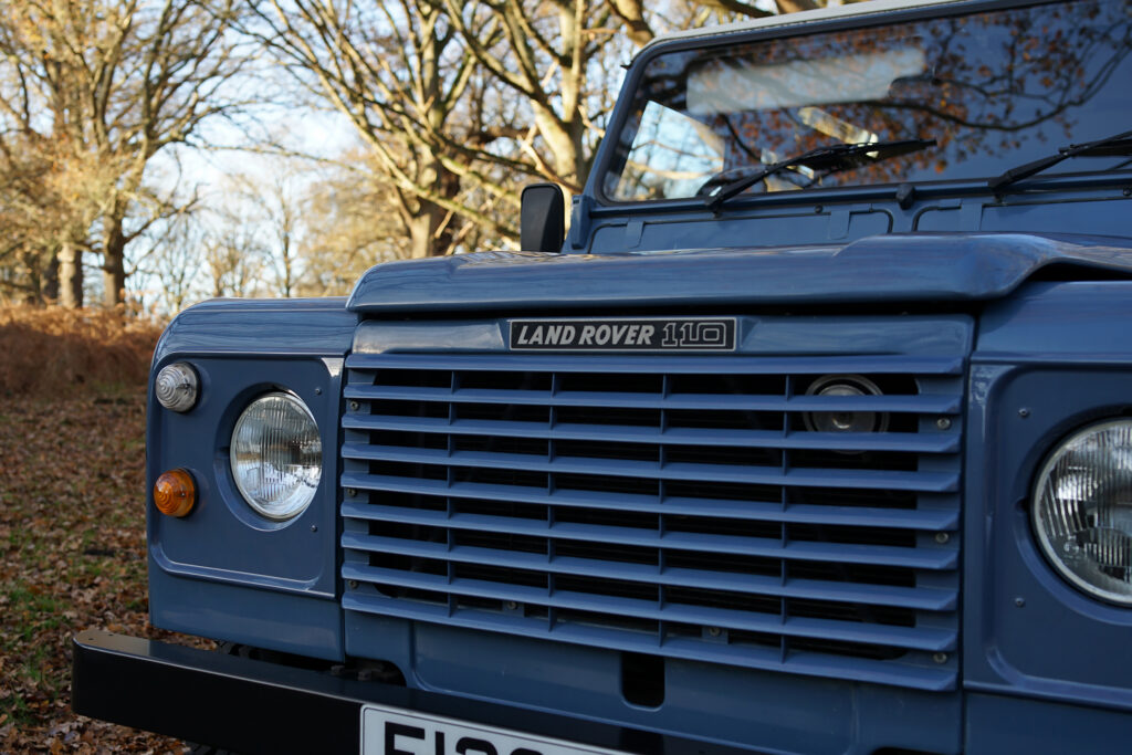 Close-up of a blue Land Rover 110 front grille and headlights with autumn trees in the background