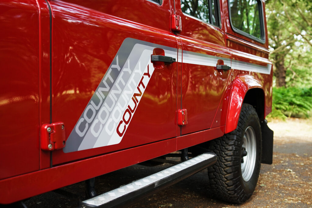 Red Land Rover with County branding on the side panel, parked on a forest road.