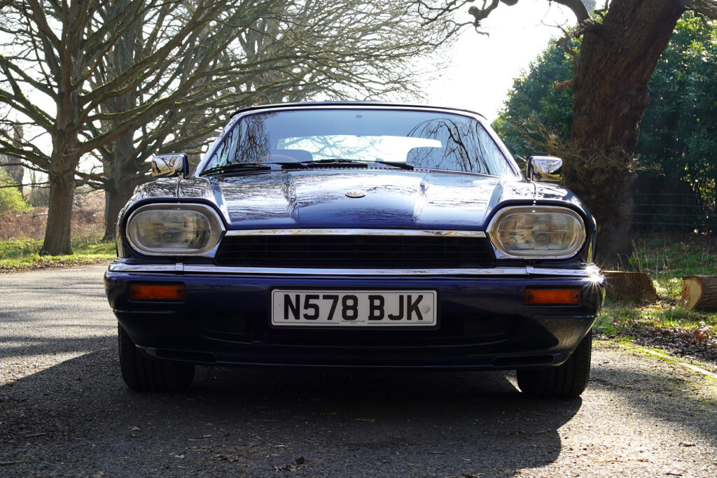 Front view of a classic dark blue Jaguar XJ-S car parked on a tree-lined driveway in natural daylight