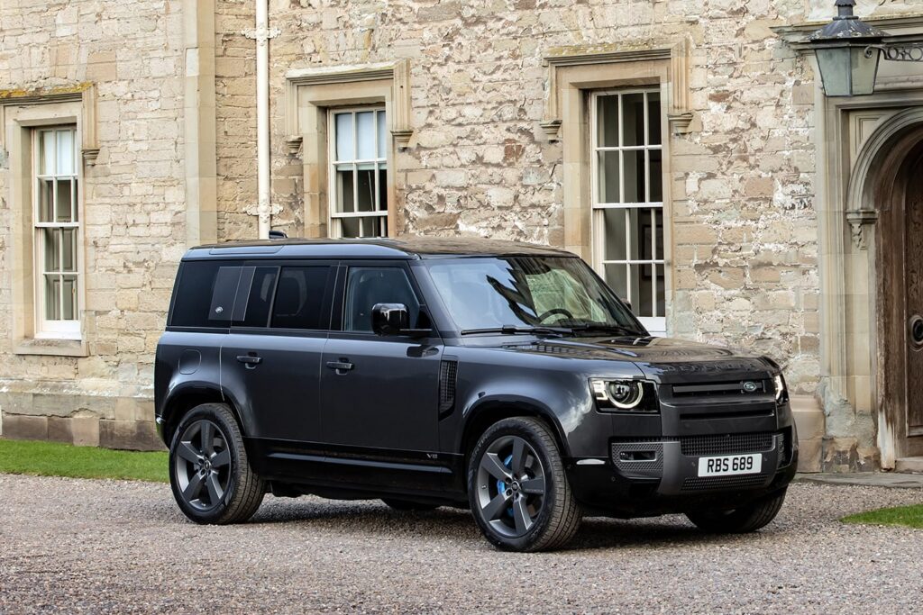 Black Land Rover Defender V8 SUV parked on gravel in front of a stone building with windows and wooden door