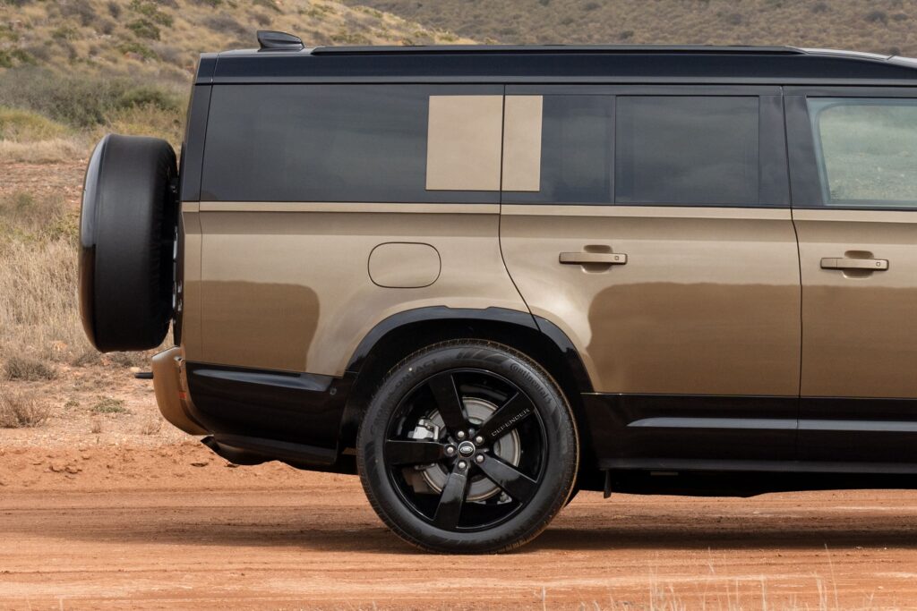 Brown and black Land Rover Defender showing rear side door, wheel with Defender branding, and spare tyre cover in a desert environment