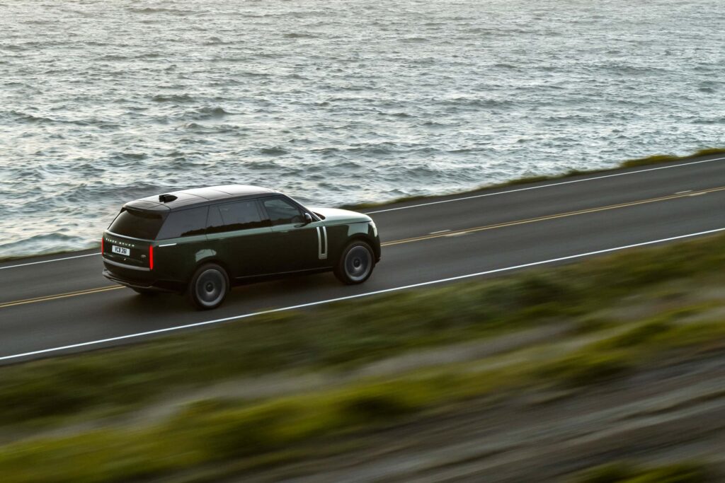 Dark green Range Rover SUV driving on a coastal road beside the ocean, showing the vehicle's side and rear view in motion.