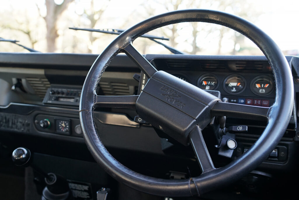 Close-up of a Land Rover classic steering wheel and dashboard showing gauges and gear stick.