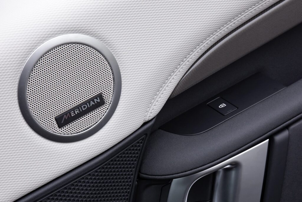 Round Meridian speaker with silver grille and black trim set into white textured interior car door panel with window control button and door handle
