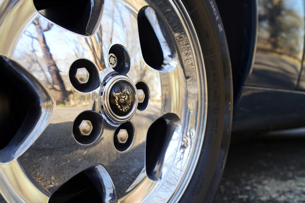 Close-up of a shiny chrome car wheel with a jaguar emblem reflecting trees and pavement, highlighting vehicle detail.