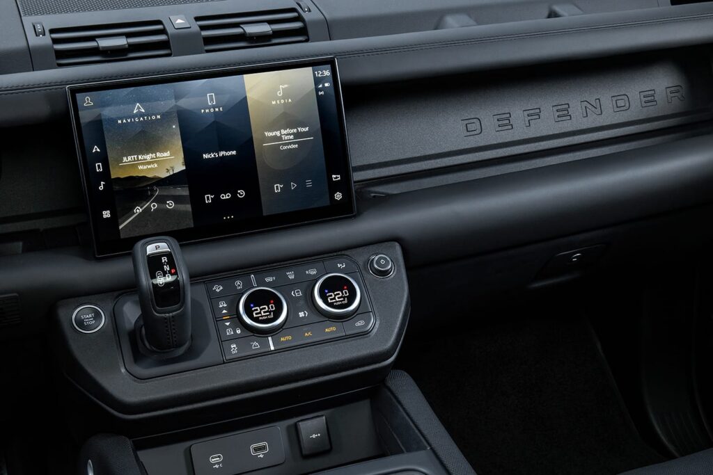 Central touchscreen displaying navigation, phone, and media in a Defender car interior with gear shift and temperature controls.