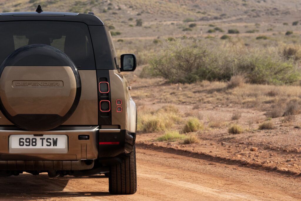 Rear view of a tan Land Rover Defender parked on a dusty dirt road in an arid, bushy landscape.