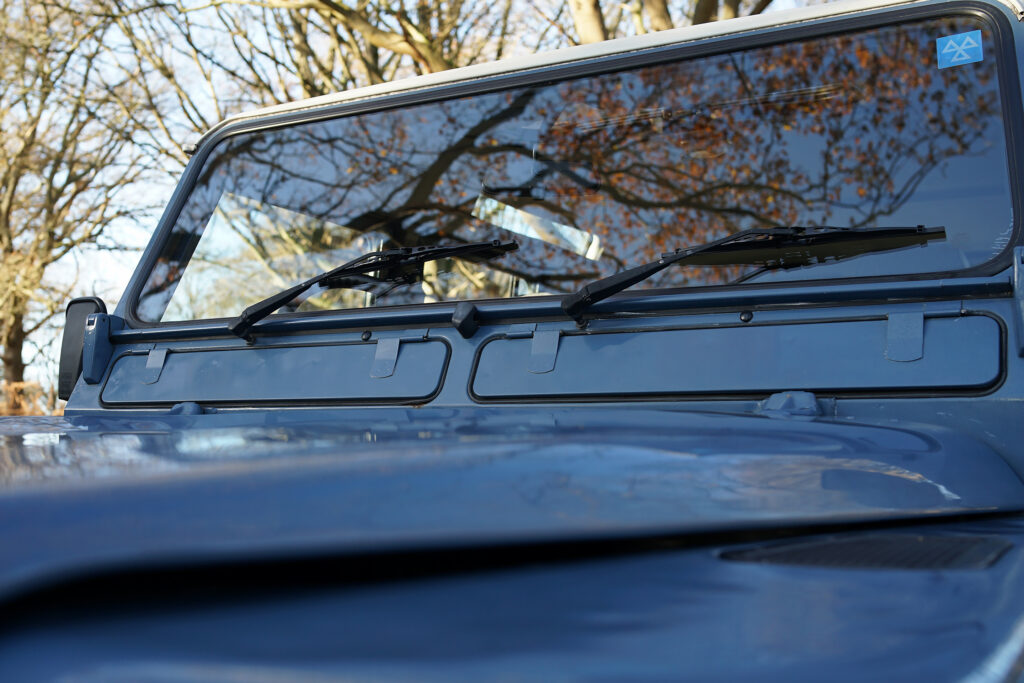 Close-up of a blue vehicle's windshield with two black wipers and reflections of bare trees, highlighting the windshield for automotive care.