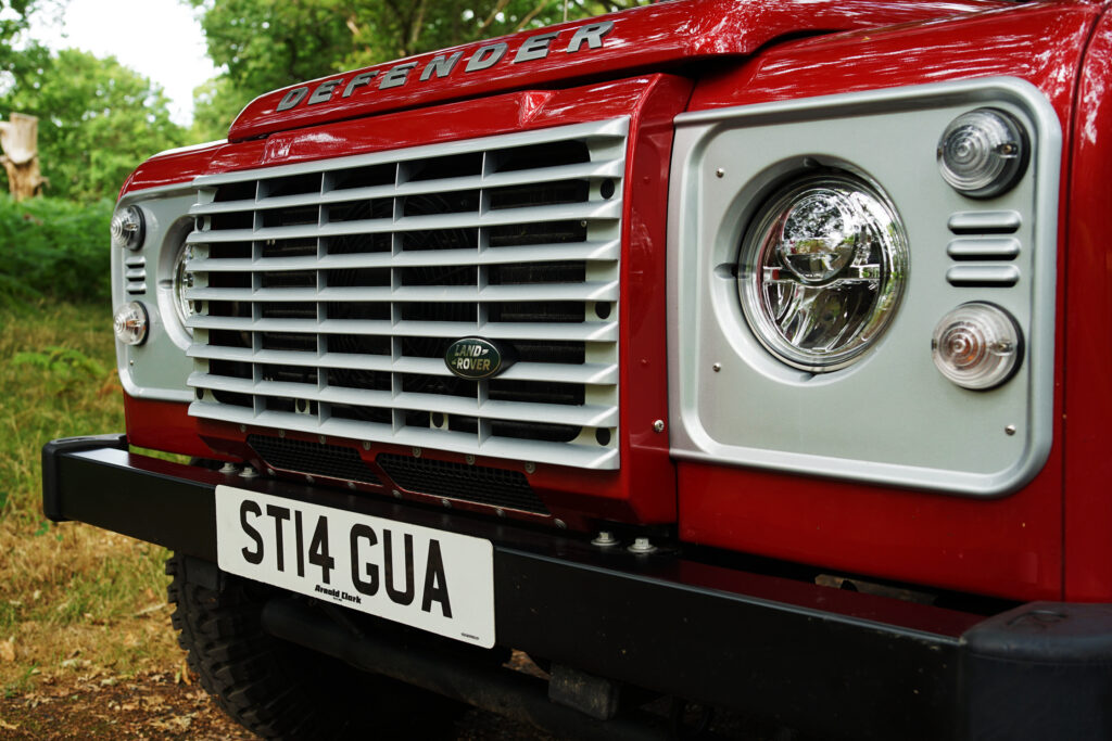 Front view of a red Land Rover Defender showing the grille, headlight, and number plate in a wooded area