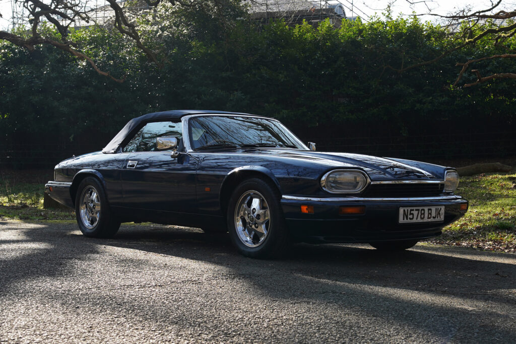 Dark blue classic convertible car with a soft top parked on a sunlit quiet road with trees in the background