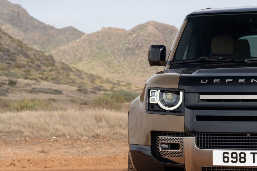 Front half of a brown Land Rover Defender with headlights on against a backdrop of arid hills and grassland
