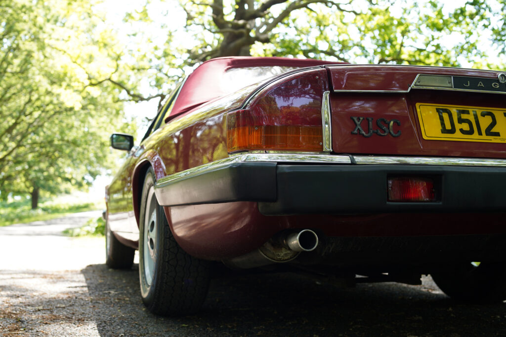 Rear view of a classic maroon Jaguar XJ-SC convertible parked on a leafy road.
