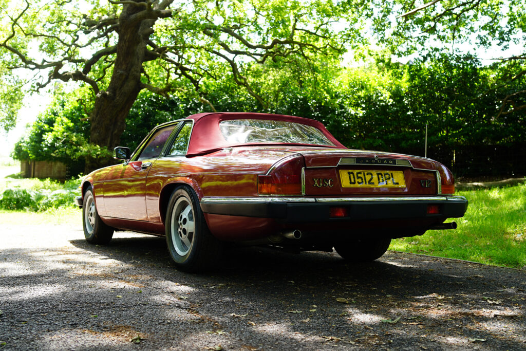 Red vintage Jaguar XJ-SC V12 convertible parked on a shaded driveway surrounded by green trees and sunlight