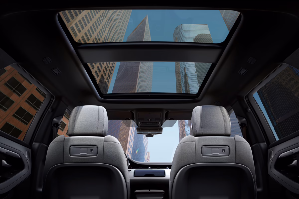 View of a luxurious car interior featuring grey front seats and a panoramic sunroof showing skyscrapers above, highlighting premium design and openness.